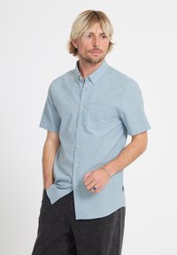 Homme blond portant une chemise à manches courtes bleu clair à boutons avec poche poitrine, pantalon foncé et une bague à la main gauche, debout sur un fond blanc.