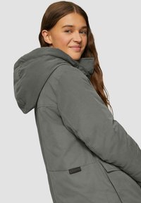 Veste grise matelassée avec capuche, en tissu texturé et poches latérales. Comprend une étiquette de logo près de l'ourlet.