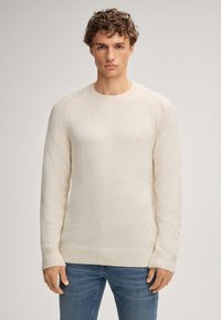 Giovane con capelli ricci che indossa un maglione di maglia crema e jeans blu, in piedi contro uno sfondo chiaro e uniforme, rivolto in avanti.