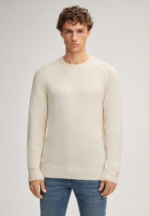 Junger Mann mit lockigem Haar, der einen cremefarbenen Strickpullover und blaue Jeans trägt, vor einem einfarbigen, hellen Hintergrund stehend und nach vorne blickend.