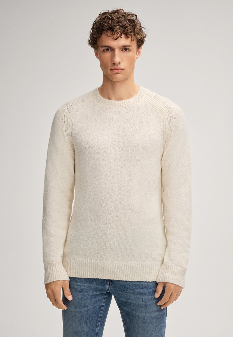 Giovane con capelli ricci che indossa un maglione di maglia crema e jeans blu, in piedi contro uno sfondo chiaro e uniforme, rivolto in avanti.