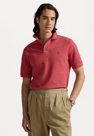 Rotes Polohemd mit Kragen, kurzen Ärmeln und kleinem blauen Logo auf der linken Brust; kombiniert mit hellen, khakifarbenen, gefalteten Hosen.