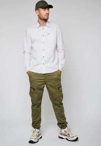 Wit blouse met lange mouwen en blauw ruitpatroon, bruine knopen; olijfkleurige cargobroek met zijzakken; wit en rode sneakers; groene pet.