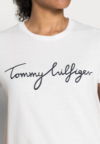 Bílá bavlněná tričko s výrazným černým kurzívním logem, které zní "Tommy Hilfiger." Klasický střih s kulatým výstřihem. Měkká textura.