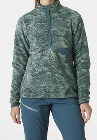 Fleece pullover in teal met golvend patroon, halfgemakkelijke sluiting en een borstzak. Gestyled met blauwe broek met een ritszak.