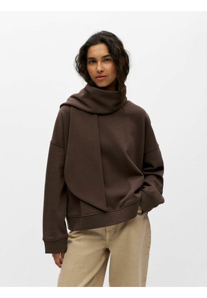 Femme aux cheveux bouclés et foncés porte un sweat-shirt marron ample avec un détail d'écharpe attaché et un pantalon beige, debout avec une main dans la poche.
