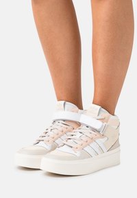 Höga skor i vitt och ljusrosa, med en kombination av läder och mocka, snörning framtill och justerbart rem för stöd.