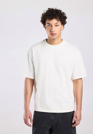 Young man with curly dark hair wearing a plain white short-sleeve T-shirt and black pants, standing against a light background.