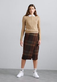 Beige cable knit sweater, white collared shirt underneath, brown plaid wrap skirt, white sneakers with light gray socks, standing on gray floor.