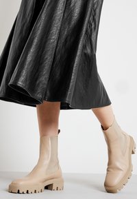 Beige ankelboots med elastiska sidopaneler och kraftiga sulor, kombinerade med en svart, glänsande, flödande kjol. Slät lädertextur.