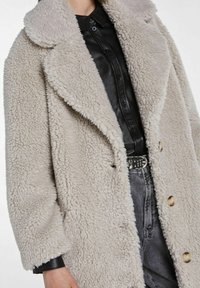 Personne portant un manteau beige à texture duveteuse sur une chemise en cuir noire boutonnée rentrée dans un jean gris avec une ceinture noire cloutée.