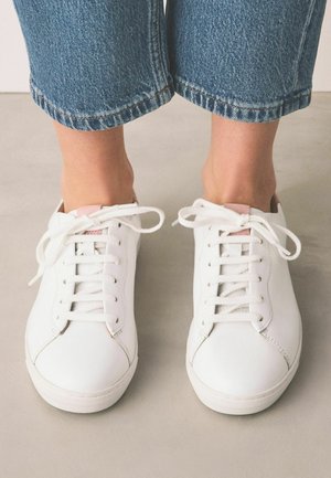 Person iført hvide snørebåndssneakers og korte blå jeans, stående på en glat, lysfarvet gulv.