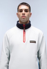 Hvid fleece-pullover med høj krave, rød lynlås og marineblå detaljer. Har et logo-patch på brystet og sømdetaljer i siden.