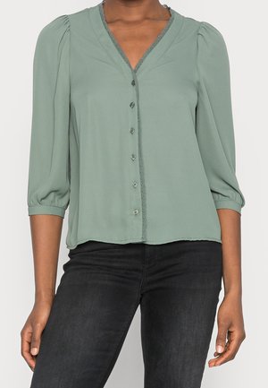 Femme portant un chemisier vert sauge avec boutons, manches ballon trois-quarts et pantalon noir ajusté, debout devant un fond clair.