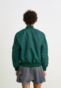 Zeleni bomber jacket s rebrastim ovratnikom, elastičnim manžetama i donjim rubom, s glatkom, sjajnom teksturom i blago podstavljenim dizajnom.