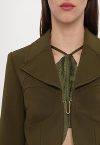 Jachetă ajustată de culoare verde măslinie, cu un guler lat, închidere cu fermoar în față și un accent de țesătură verde texturată la decolteu.