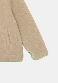 Beige Fleecejacke mit einer weichen, strukturierten Oberfläche, die grüne Akzente und eine Tasche auf der Vorderseite aufweist. Abgerundete Bündchen verstärken das Design.