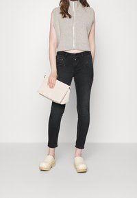 Suéter sin mangas de punto con cierre de cremallera, combinado con jeans ajustados oscuros y zuecos claros, sosteniendo un bolso de mano color crema.