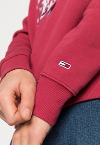 Detailní záběr na ruce, které upravují manžetu červeného svetru s malým obdélníkovým logem, v kombinaci s modrými džíny.