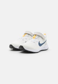 Fehér sportcipők hálós felsőrésszel, sötétkék swoosh logóval, sárga kiemeléssel a nyelvén, szürke pánttal és fekete gumitalppal.