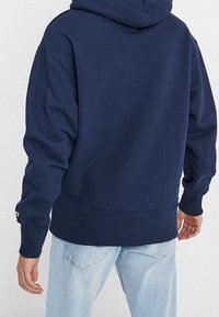 Felpa blu navy con tasca frontale a marsupio, polsini e orlo a coste, e cappuccio con coulisse. Il tessuto appare morbido e leggermente testurizzato.