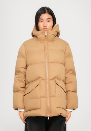 Woman wearing a camel-colored puffer jacket with large front pockets and a high collar, standing against a plain white background.