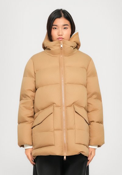 Vrouw in een beige pufferjack met grote zakken aan de voorkant en een hoge kraag, staande tegen een effen witte achtergrond.