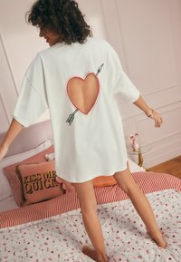 Witte oversized t-shirt met een hartvormige uitsnijding op de rug, voorzien van rode stiksels en een pijlontwerp. Zachte textuur, relaxte pasvorm.