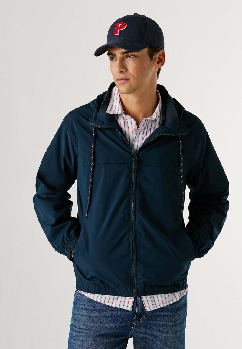 Veste coupe-vent bleu marine avec capuche, poignets élastiques, cordons de serrage et deux poches, assortie à une chemise rayée et un jean foncé.