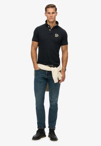 Schwarzes Polohemd mit Kragen und Logo, kombiniert mit blauen Jeans. Ein cremefarbener Pullover ist um die Taille gebunden. Schwarze Schuhe runden das Outfit ab.