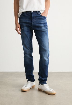 Jeanși din denim de culoare albastru închis, cu croială slim, având decolorări subtile și efect de uzură. Asortați cu adidași albi, stând pe un podea neutră.