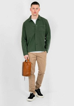 Green button-up shirt with a chest pocket, beige pants, black sneakers, and a brown leather bag with blue and white zipper accents.