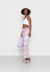 Jeans lilás com padrão tie-dye e corte descontraído, combinados com um top cropped branco e acessórios lilás a condizer, em fundo neutro.