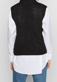 Schwarzer gestrickter ärmelloser Pullover mit hohem Kragen und strukturiertem Muster, getragen über einem weißen Hemd. Blaue Jeans sind ebenfalls sichtbar.