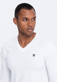 Weißes langärmliges V-Ausschnitt-T-Shirt mit einem kleinen schwarzen Logo auf der linken Brust. Weicher Stoff mit glatter Textur, taillierter Schnitt.