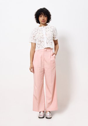 Top court en dentelle blanche avec des bords festonnés, associé à un pantalon large rose pâle avec des plis avant et des poches latérales, porté avec des mocassins argentés.