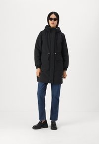 Parka noire à capuche avec fermeture éclair, deux poches avant, cordons de serrage ajustables et poignets élastiques, associée à un jean foncé et des chaussures noires.