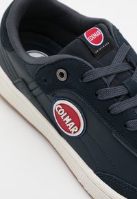 Baskets bleu marine avec tige en cuir, accents gris texturés, logo rond rouge et blanc, lacets gris et semelle en caoutchouc avec un motif texturé.