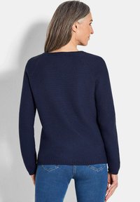 Frau mit langen grauen Haaren, die einen marineblauen strukturierten Pullover und eine Jeans trägt, mit dem Rücken zur Kamera und mit leicht nach rechts gedrehtem Kopf.