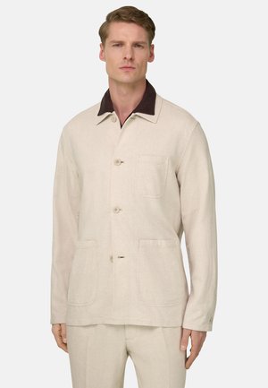 Uomo che indossa una giacca beige con bottoni, tre tasche frontali e un colletto marrone scuro, in piedi contro uno sfondo semplice.