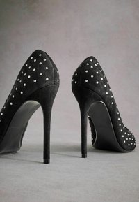Mustat mokkanahka korkokengät, joissa on terävä kärki ja hoikka stiletto-muotoilu, koristeltu pienillä valkoisilla niiteillä koko päällisen pinnalla.