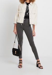 Chaqueta de mezclilla blanca, camiseta gráfica negra, jeans skinny gris de cintura alta, bolso negro y tacones negros con tiras.