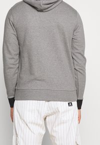 Sweat à capuche gris avec une texture lisse, des poignets ajustés et une capuche avec cordon central. Porté avec un pantalon cargo rayé blanc.
