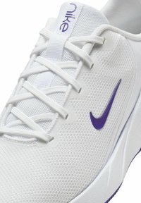 Zapatilla de correr blanca de malla con una superficie texturizada, que presenta un logo de Nike púrpura y acentos, cordones planos y una suela blanca acolchada.