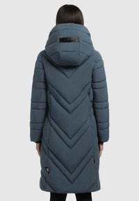 Pitkä, muhkea, tummansinisestä chevron-tikkauksesta valmistettu takki, jossa on suuri huppu ja logo takana. Ominaisuuksiin kuuluu vaakasaumastus ja sivutaskut.