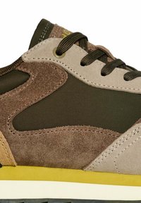 Detalle de zapatilla de ante marrón y tejido verde con cordones oscuros, acentos amarillos en la suela y costuras de punto, que presentan un diseño de ola.