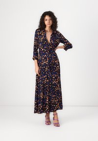 Maxi dress in black fabric featuring a multicolored leaf pattern in orange and blue. Long sleeves and a V-neckline. Paired with purple sandals.