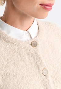Flauschige cremefarbene Strickjacke mit strukturierter Oberfläche, ausgestattet mit einem runden, goldakzentuierten Knopf und einem weißen Hemd darunter.