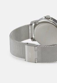 Correa de reloj de malla plateada con un cierre de acero inoxidable, que presenta un logo grabado. Caja de reloj redonda visible, que muestra un diseño sencillo.