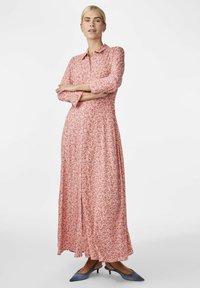 Robe maxi florale rose avec un design à boutons, des manches longues et une silhouette fluide. Portée avec des talons aiguilles bleus.
